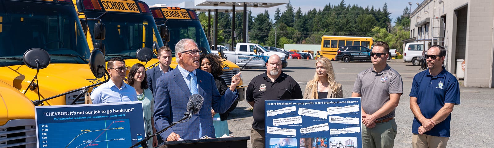 Photo of Gov. Inslee standing outdoors at a podium with a small group of people behind him. Behind the group are new electric school buses, and next to the podium are two posters — one showing increasing oil company profits and another showing a collage of headlines about oil company profits and climate-related weather events and disasters.