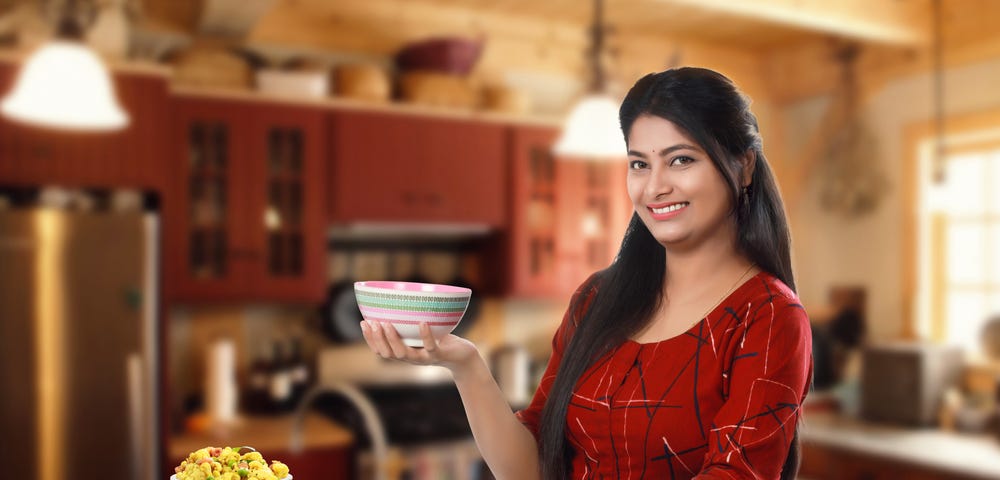 Smiling Indian girl holding a bowl sitting in relax mood beside group of traditional Indian dishes and looking at the camera.