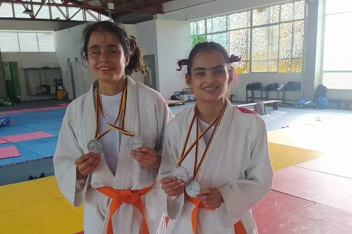 Andrea Orozco Escárraga, junto a una compañera de equipo a su izquierda. Ambas visten kimonos de judo blancos con cinturones de color naranja. Cada una luce dos medallas de competición. Ambas sonríen a la cámara.
