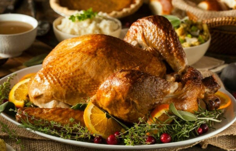 A roast turkey on a platter with herbal garnishes, with bowls of mashed potatoes, gravy, rolls, and pie on a table.