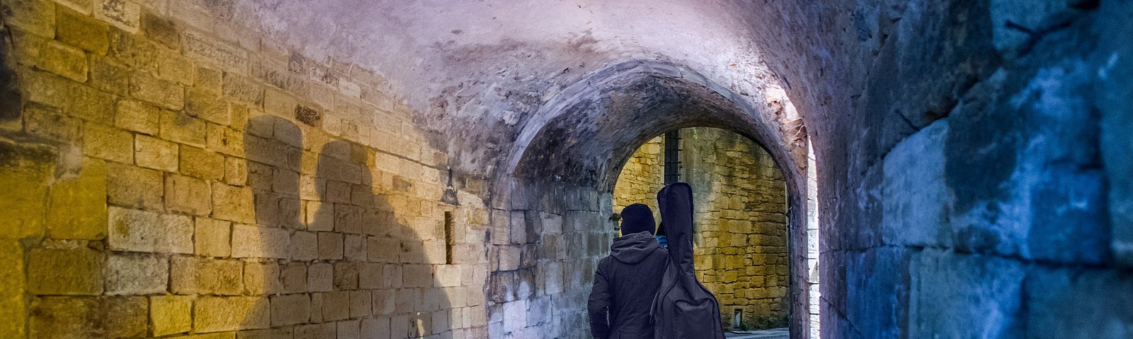 A tunnel between stone walls, shaded in yellow-orange and blue. A person clothed in black walks away on the cobblestones, carrying a guitar in a soft case over one shoulder.