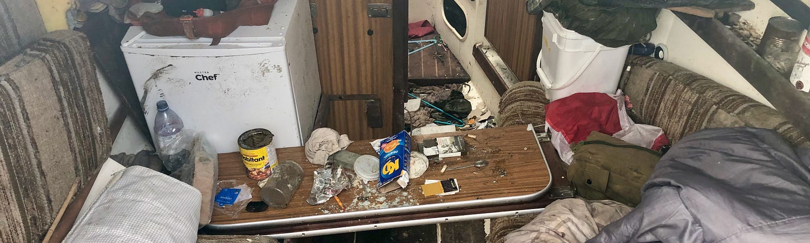 A very untidy scene. The camera looks down into a boat. A small table is covered in debris, a fridge is in the corner and bedding litters one side.