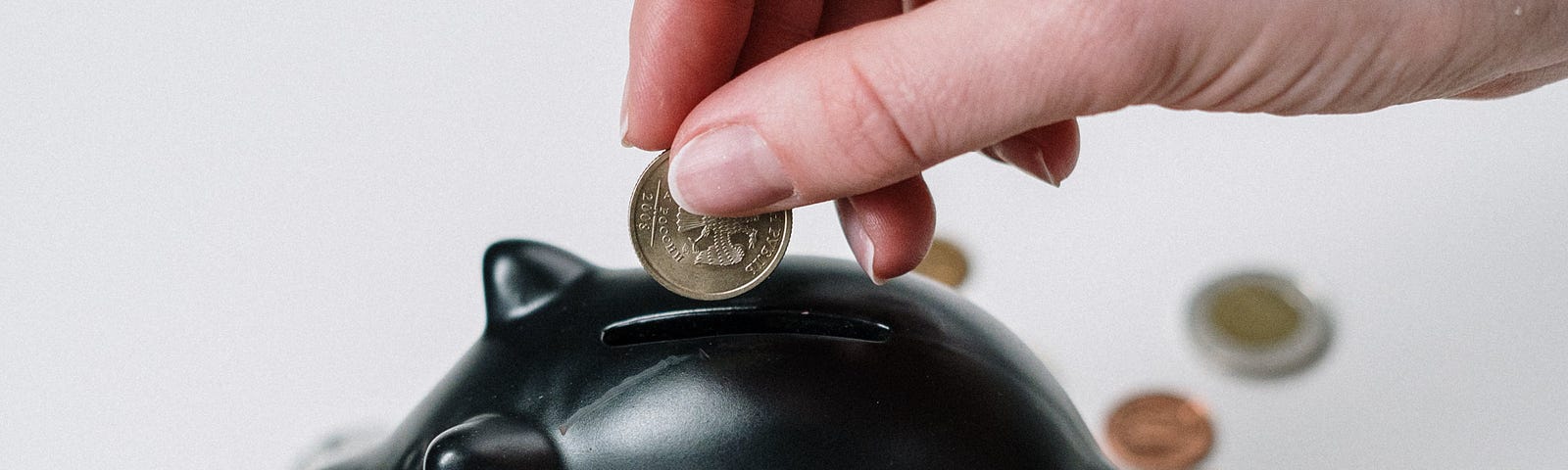 Hand placing a coin into a black pottery piggy bank that is standing on other coins.