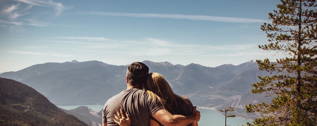 A rear view of a man and a woman sitting on a bench on top of a tall hill. They are looking out over a large lake with mountains in the distance. They each have an arm around the other’s back in a loving embrace, and she is resting her head on his shoulder.