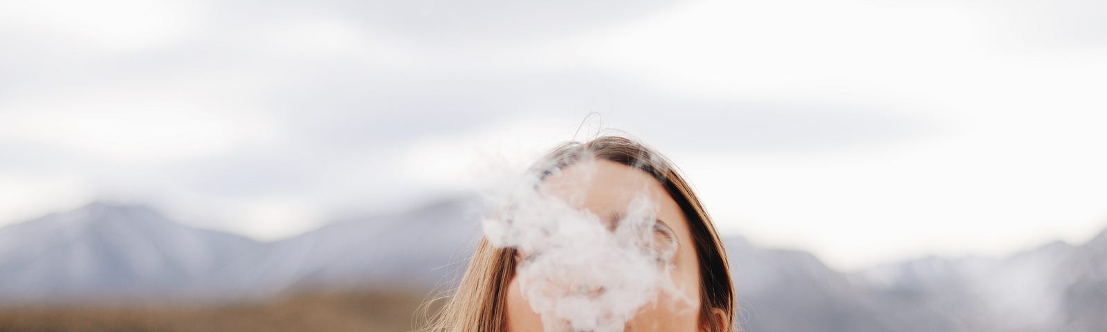 Woman blows a puff of smoke into the air while standing in front of a mountain landscape.