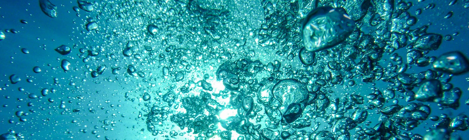 Photo: looking up through bubbles toward the surface of a large body of water. Water is blue and can see sunlight.
