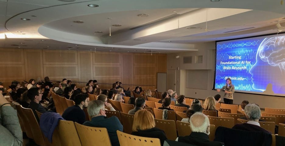 A woman speaks to a packed room of people. A large screen behind her displays a blue slide with the words, “Starting foundational AI for brain research.”
