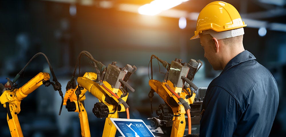 Worker in a hard hat and jumpsuit looking at a laptop that’s controlling a machine