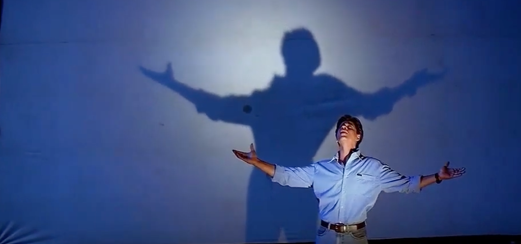 Actor Shahrukh Khan stands in front of a projector screen with his shadow on it spreading his arms and looking towards the sky