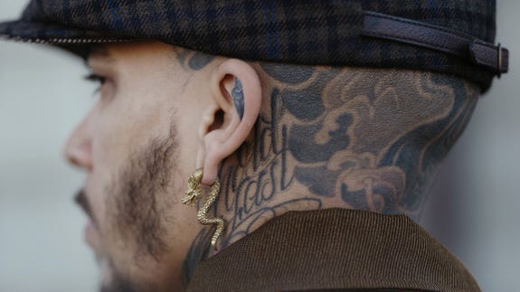 Close-up of man with gold dragon earring, head tattoos and a cap