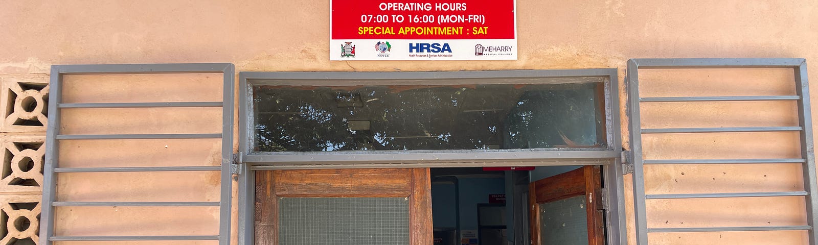 Esnart is wearing a bright pink blouse, and is standing outside of the maternal and child health ward, which is a beige pink building with a bright red sign.