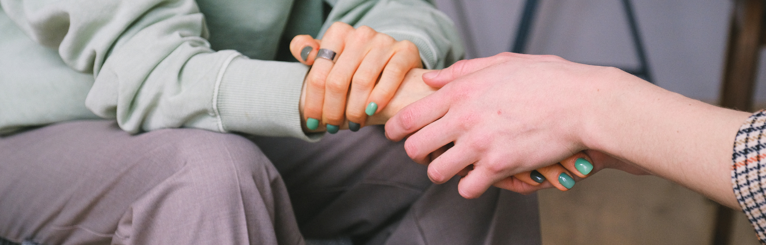 two people sitting across from each other, a hold holding another hand, the second hand’s fingernails are visible and green from A New Mental Health Toolkit From To Write Love On Her Arms (TWLOHA) by Gem Blackthorn