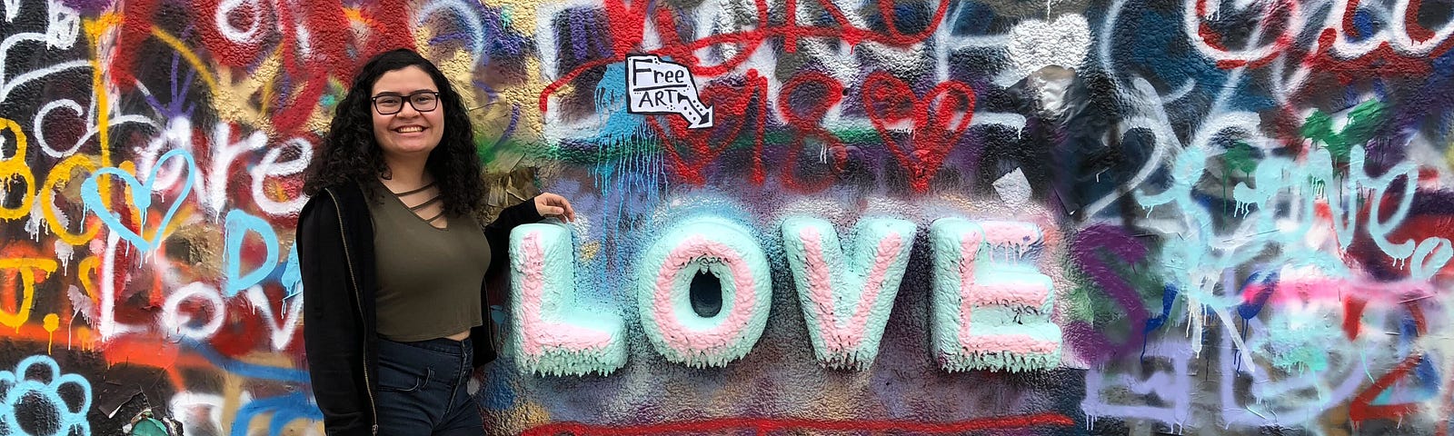Miriam, the writer of this blog post, is posing next to a wall of graffiti art, specifically in front of a large 3D piece of art that says, “LOVE”