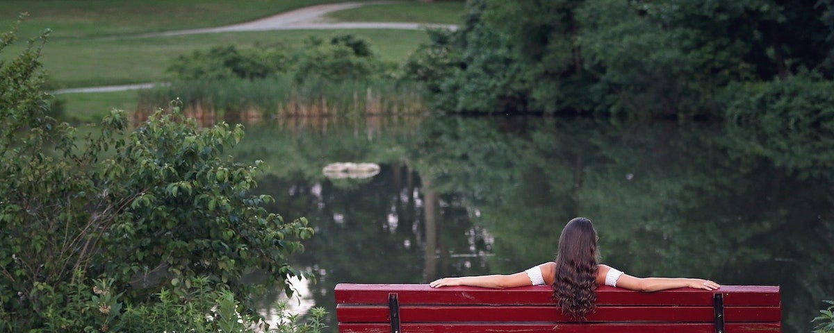 A rear view of a woman sitting alone on a park bench, looking out over a lake. She has her arms stretched out along the back of the bench.