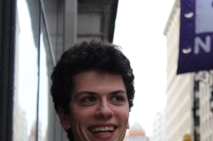 Man with brown hair standing on the sidewalk with an NYU flag behind him.