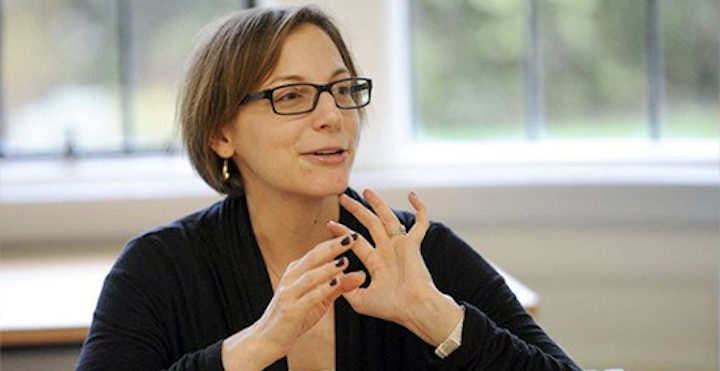 A woman with short hair and glasses speaks to an unseen person off camera.