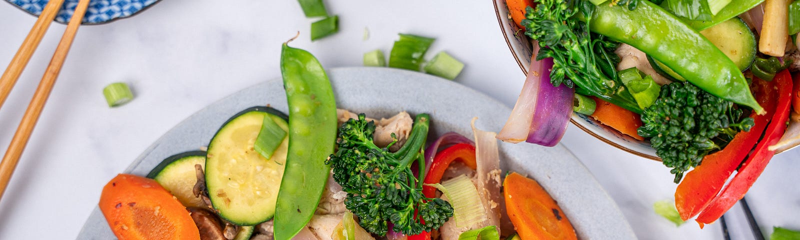 A top-down view of a plate and a bowl of stir fry with chicken, broccolini, carrots, zucchini, red peppers, mushrooms, and green onions.