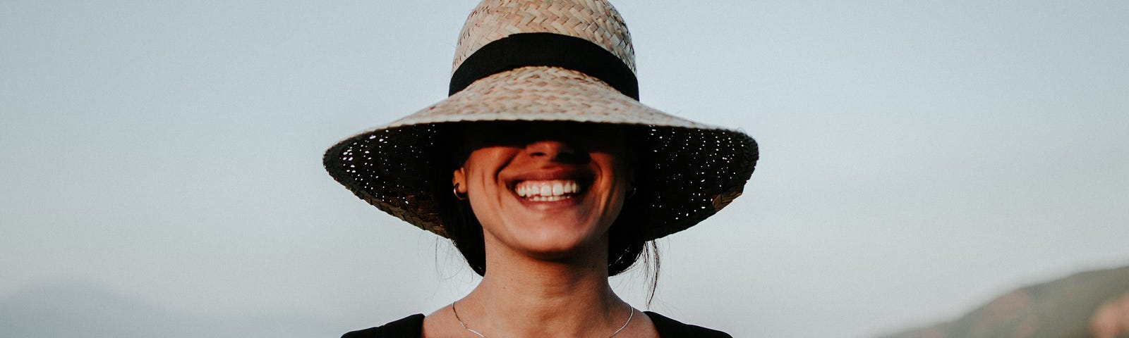 A woman smiling at the beach with a big hat covering half of her face