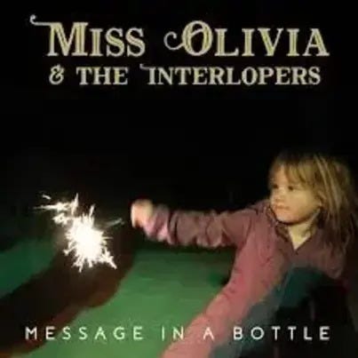 Miss Olivia & The Interlopers’ “Message in a Bottle” single cover art; little blond girl in reddish-purple jacket playing with a sparkler in the dark, band name at top and single name at bottom