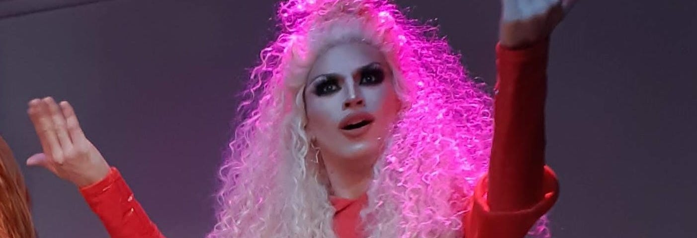 The drag queen Aquaria performs with long pink hair and makeup in a long sleeved red dress.