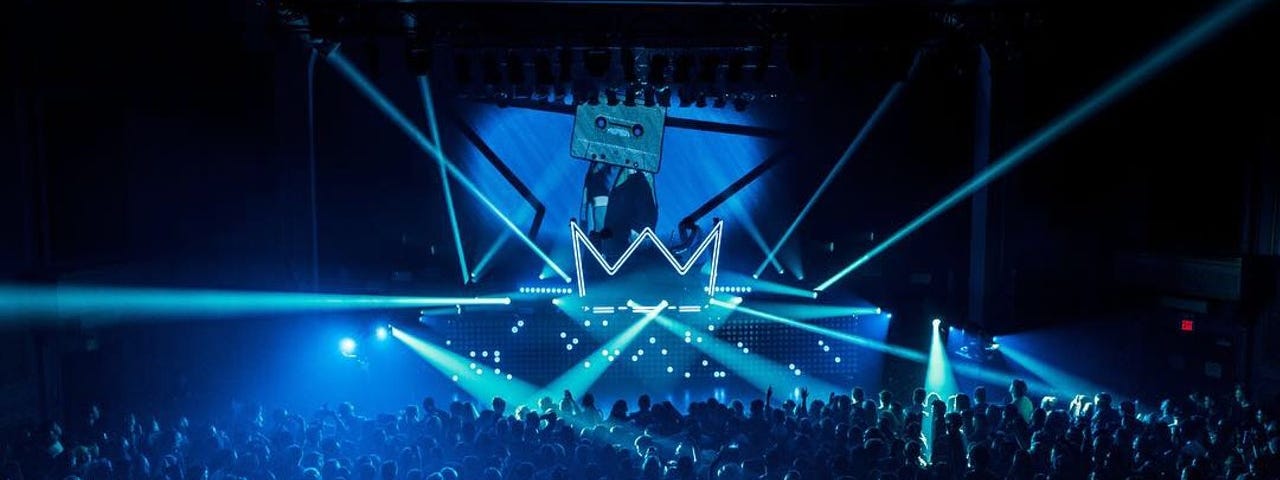 a huge stage is set with a laser show in blue and aqua. thousands of people fill the crowd in front of it.