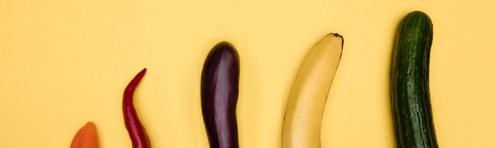 Five vegetables — two peppers, an aubergine or eggplant, a banana, and a cucumber — laid out in a row. Each is longer and/or thicker than the previous.