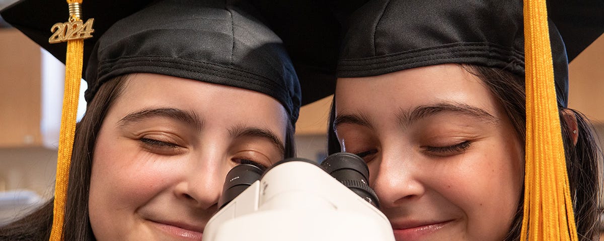 Isabella and Katelyn De Leon looks through a mircroscope while wearing their 2024 graduation caps in a biology lab on the northwest denver campus