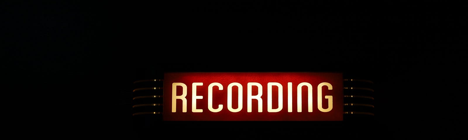 The word RECORDING in sans serif font and in all capital letters is back lit and appears on a red background with silver adornments on each side. It appears to be a sign inside a dark room.