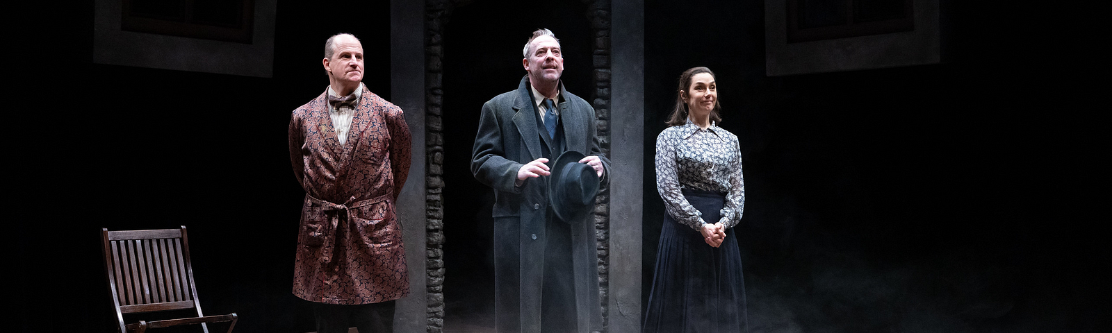 Three actors stand onstage and prepare to take a bow; the actor on the left wears a patterned smoking jacket, the one in the middle a gray suit, and the one on the right a blue skirt and floral blouse.