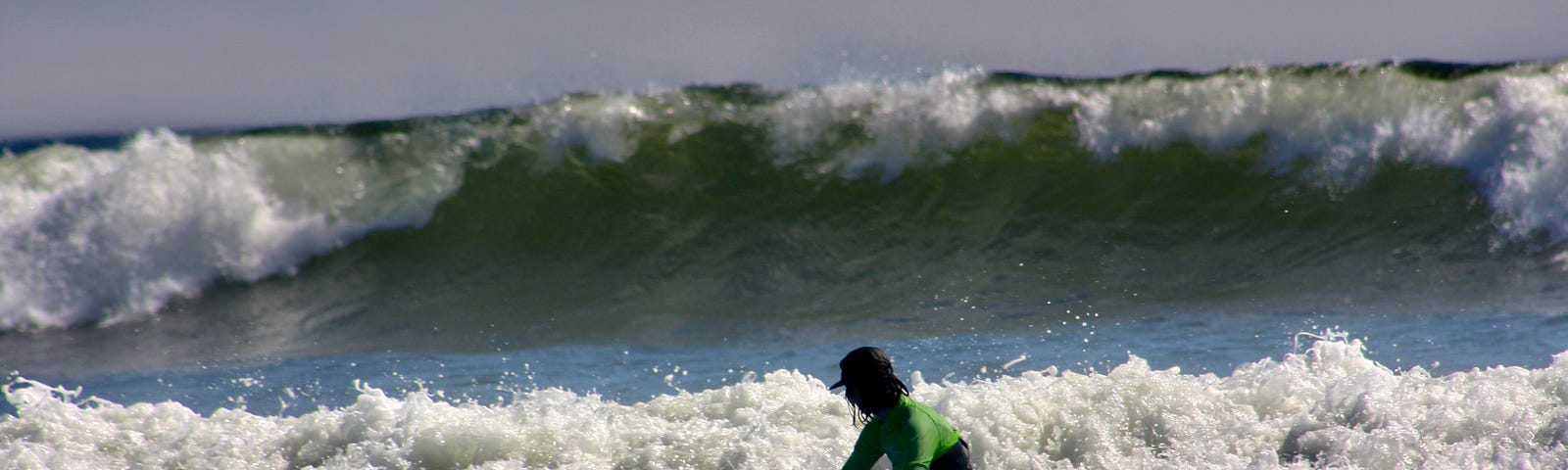 A surfer stands below a large wave that flows towards them. The water is blue and the sky is grey.