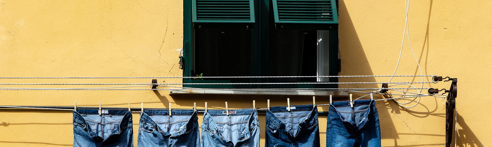 A row of denim jeans hanging to dry under a window and against a yellow wall.