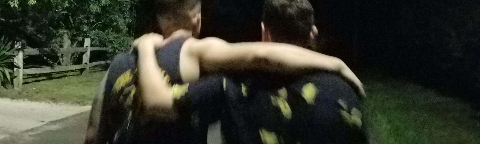 Two young men are walking with one arm on each other’s shoulders.