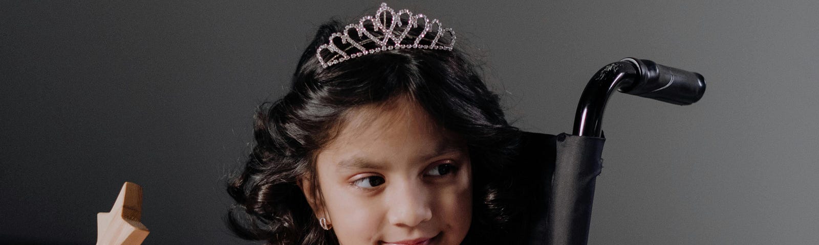 Young girl in a tiara and wand sits in her chair with a Mona Lisa smile as she looks at something off-camera.