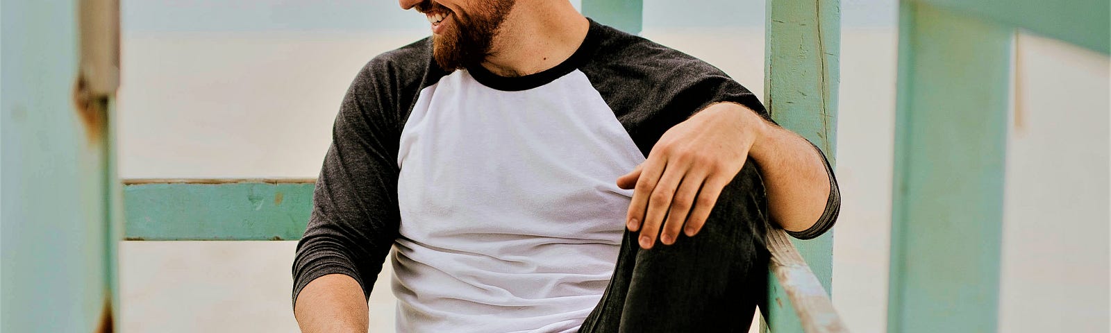 profile of smiling man with beard wearing white and black top and jeans sitting on wooden deck