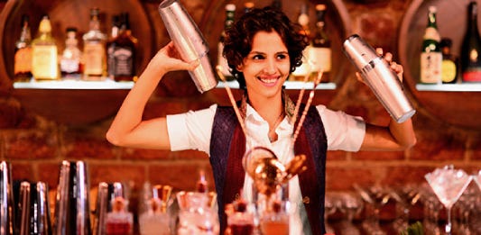 Photo of Ami Behram Shroff, renowned Flair Bartender, Mixologist and Fire Juggler, smiling and pouring drinks into a glass. She is wearing a white shirt and dark brown sleeveless jacket.