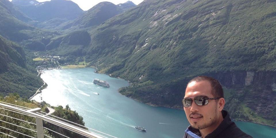 A picture of the author with a view of the blue water of the fjord and green mountains in the background