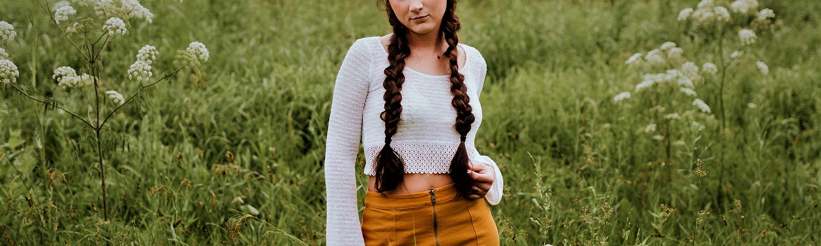 girl with long dark braids wearing long sleeve white shirt and orange skirt standing in grassy field