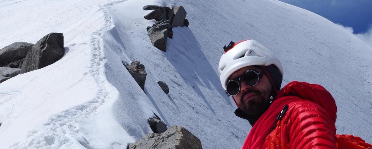 Man in red down jacket and helmet stands near the summit of a mountain.