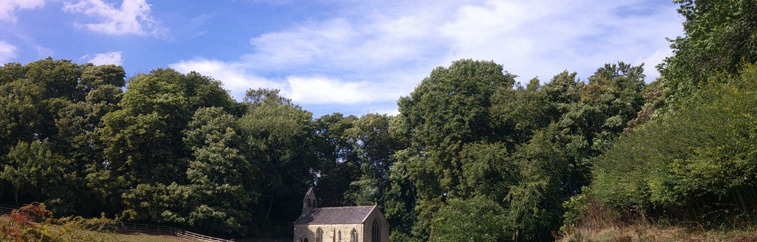 View from a hillside of St. Ethelburga’s Parish Church, Great Givendale on a bright sunny day, with trees and blue sky in the background