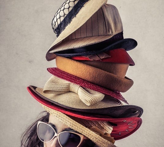 A woman wearing sunglasses looks to the side. Atop her head are a dozen different hats worn atop each other.