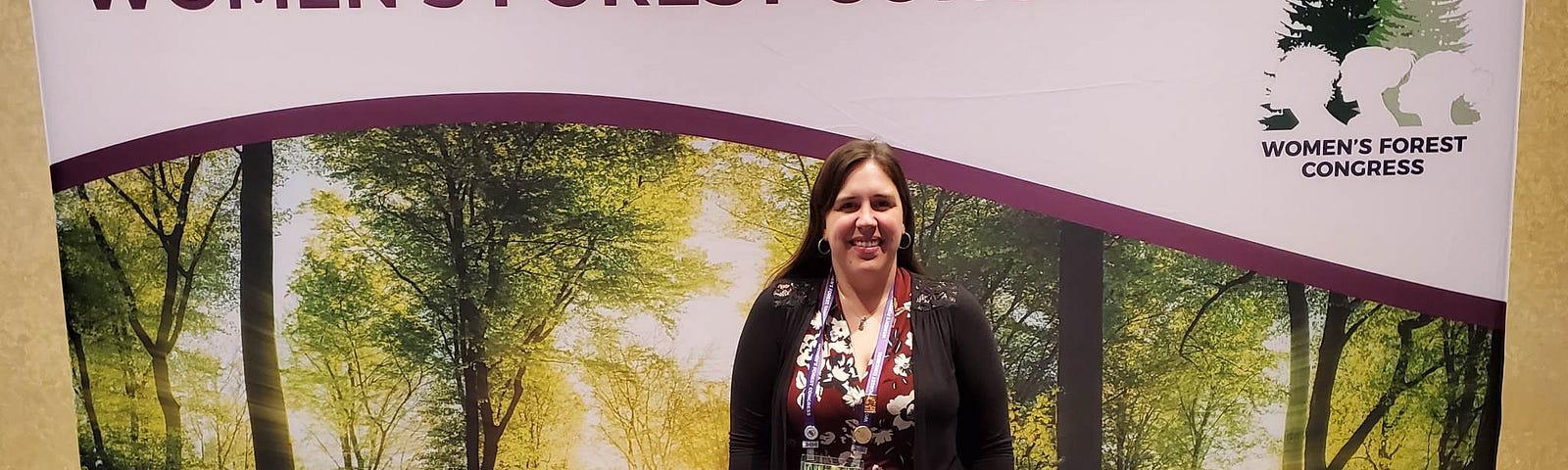 Candra Burns wearing business casual clothing in front of a sign saying Welcome to The Women’s Forest Congress with a forest background.