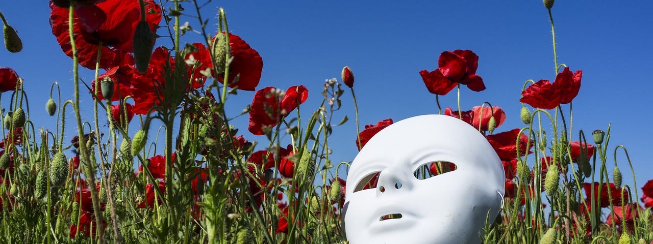 A white thespian mask lying in a field of poppies under a blue sky.