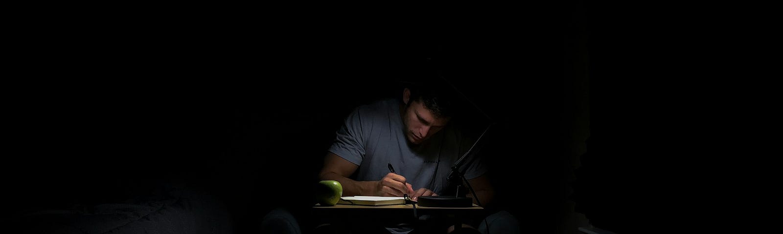 A writer is writing atop a small desk with a faint light.