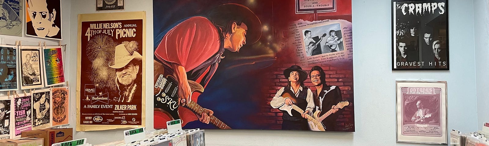 A record store with a painting of Stevie Ray Vaughan on the wall with a promo for Antone’s nightclub. There are many vinyl records in racks in front of the painting and posters for gigs on the walls.