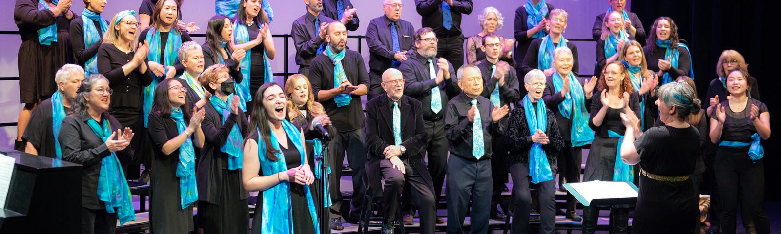 DeCoro adult choir sings on stage with a winter themed backdrop. Choir members are wearing all black clothing with blue scarves or ties.