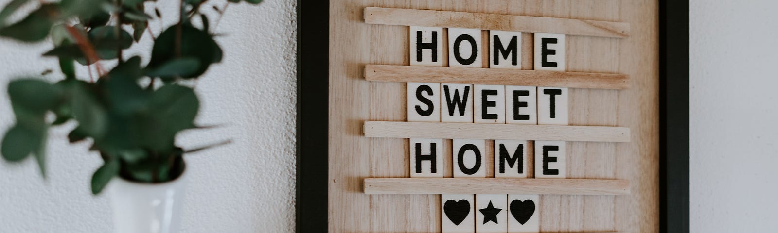 Wooden board with home sweet home written in block letters and two black hearts next to a plant