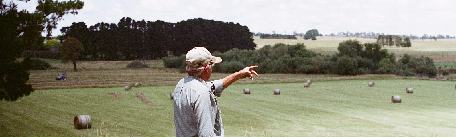 Older male farmer, standing in a pasture, pointing towards something — hay bales in the distance.