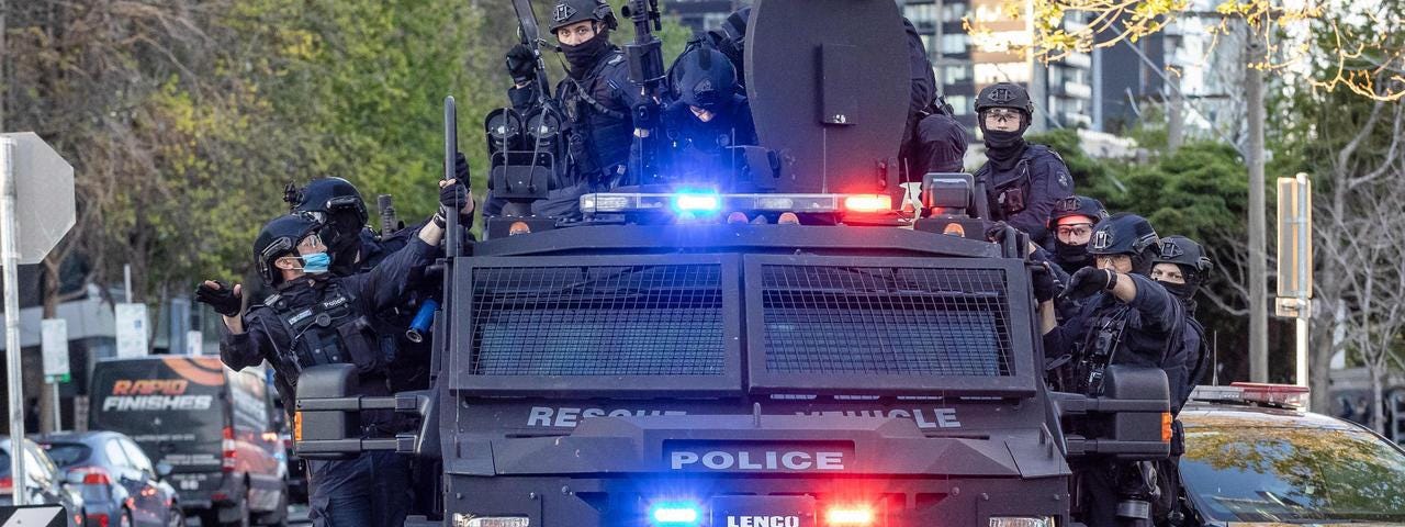 A bulky armoured vehicle labelled ‘police’ with at least ten masked cops clinging to the roof and sides, lights flashing blue and red, an array of exterior technological attachments visible on its roof