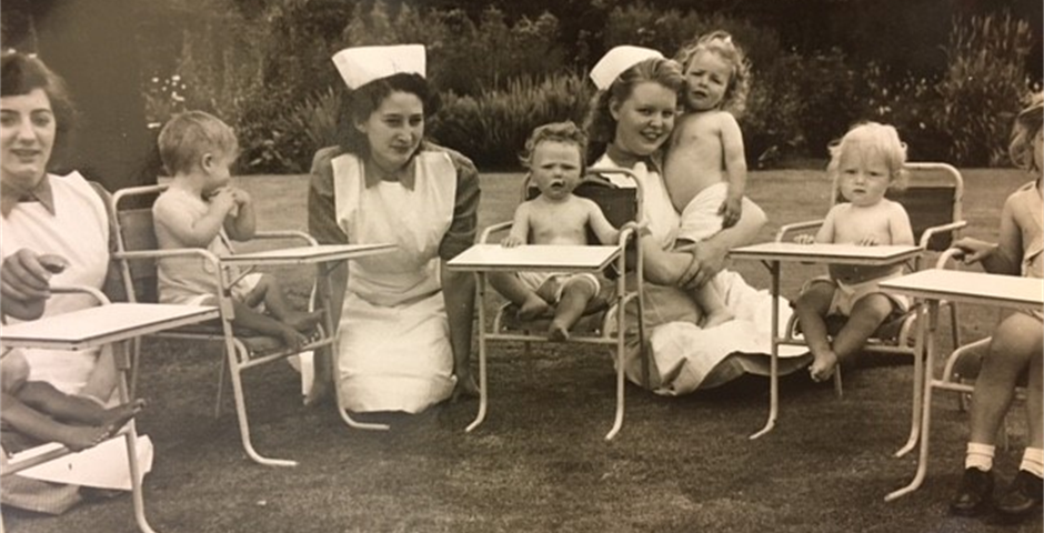 Photograph of nurses sitting next to babies in highchairs.