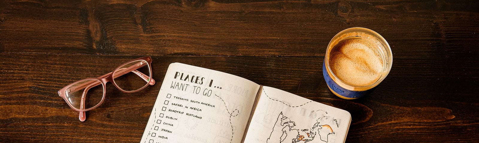 Vista superior de uma mesa de madeira com óculos, caneca com café e caderno com uma lista de lugares para viajar e um desenho de um mapa.